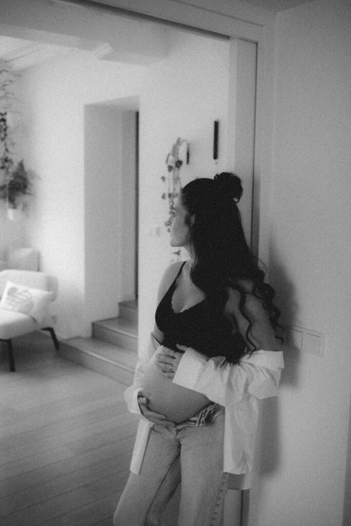 Zwangere vrouw die leunend tegen een deurpost in huis naar haar buik kijkt tijdens een zachte, winterse zwangerschapsshoot binnen.